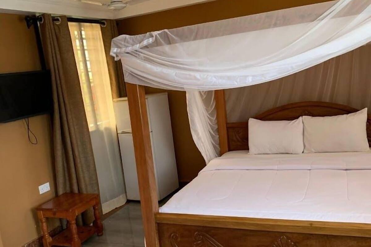 Une chambre avec un lit sous un dais, un petit tabouret en bois, un téléviseur fixé au mur et un ventilateur de plafond.