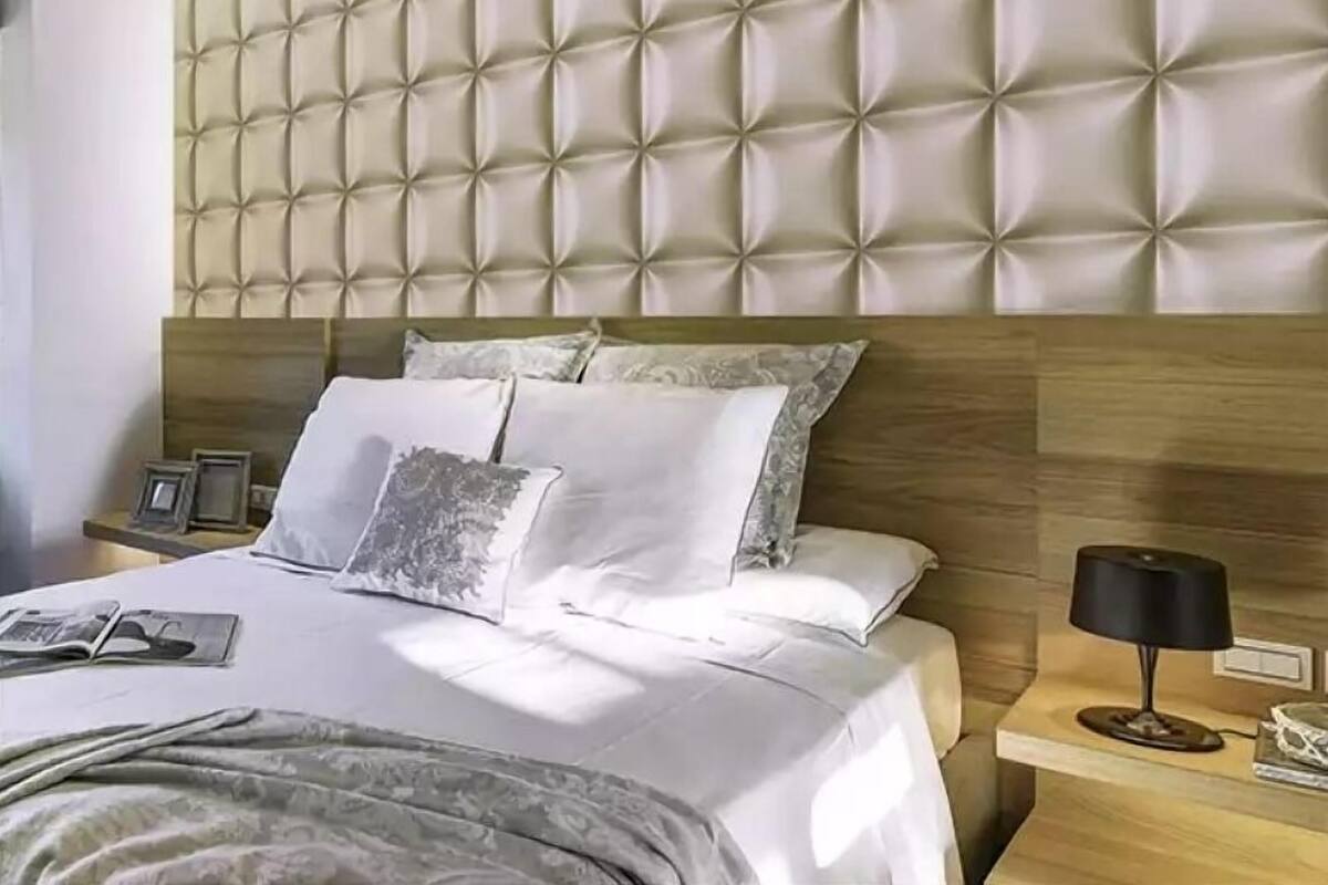 Une chambre à coucher moderne avec une tête de lit capitonnée, un lit bien fait avec du linge de lit blanc, et une table de chevet avec une lampe et un tableau encadré.