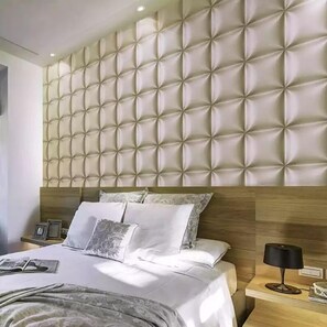 A modern bedroom with a tufted headboard, a neatly made bed with white linens, and a bedside table with a lamp and a framed picture. - Maghonyi Hotel Voi (Voi)