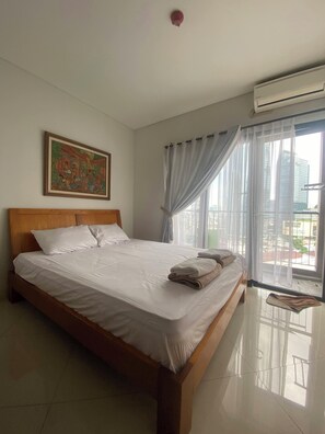 A neatly made bed with white linens and a folded towel, a wooden headboard, a painting on the wall, and a window with sheer curtains. - 2 BR Apartment in Sudirman - Kuningan Area 4-6 pax (Jakarta)