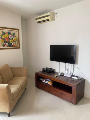 Een woonkamer met een beige bank, een houten tv-meubel met een flatscreen televisie, een wandairconditioning en een lijst met een bloemistisch schilderij.