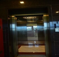 Un ascensor con interior metálico, puerta de vidrio con diseño y pared roja a la izquierda.