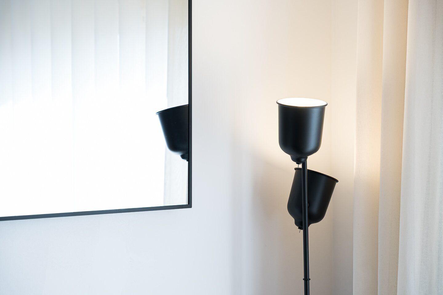 Eine moderne Stehlampe mit schwarzem Fuß und weißem Lampenschirm, platziert neben einem Fenster mit vertikalen Jalousien.