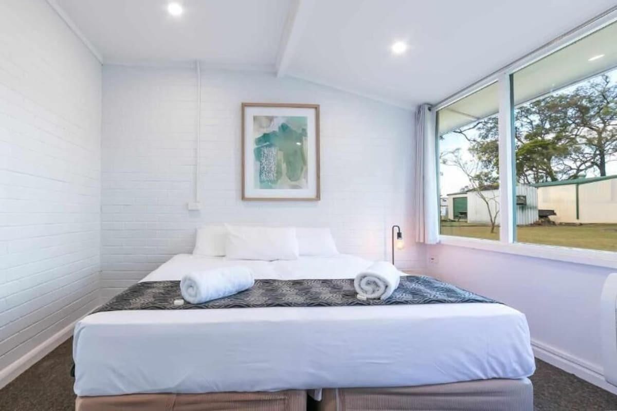Ein Hotelzimmer mit einem großen Bett, weißen Wänden, einem gerahmten Bild und einem Fenster mit Blick nach außen.