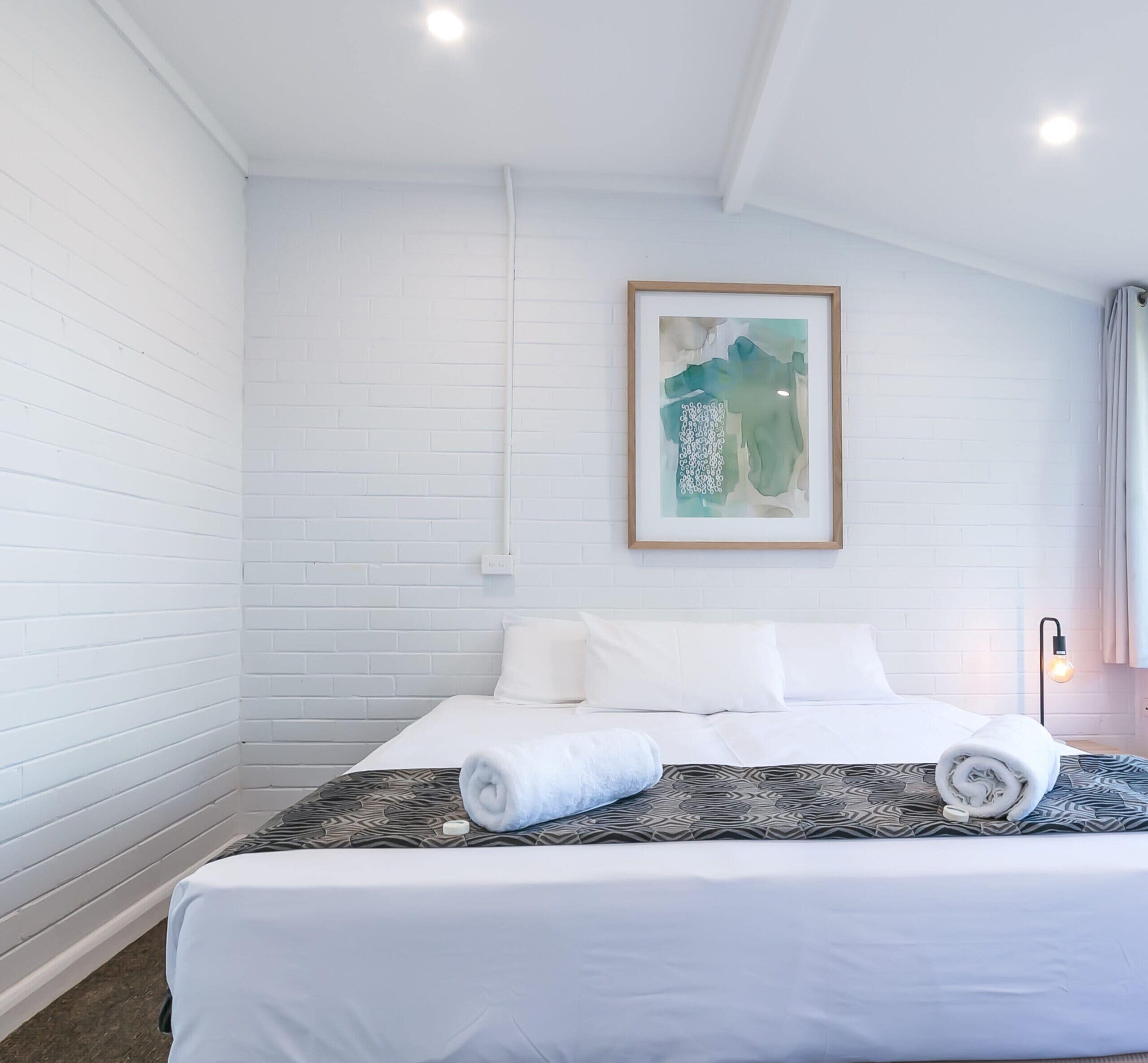 A hotel room with a bed, white walls, a framed abstract painting, and a bedside lamp.