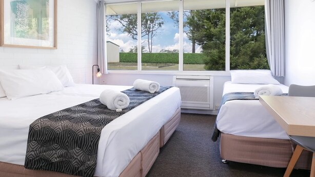 A hotel room with two beds, a desk, and a large window with a view of trees and a building. - Tahmoor Inn (Tahmoor)