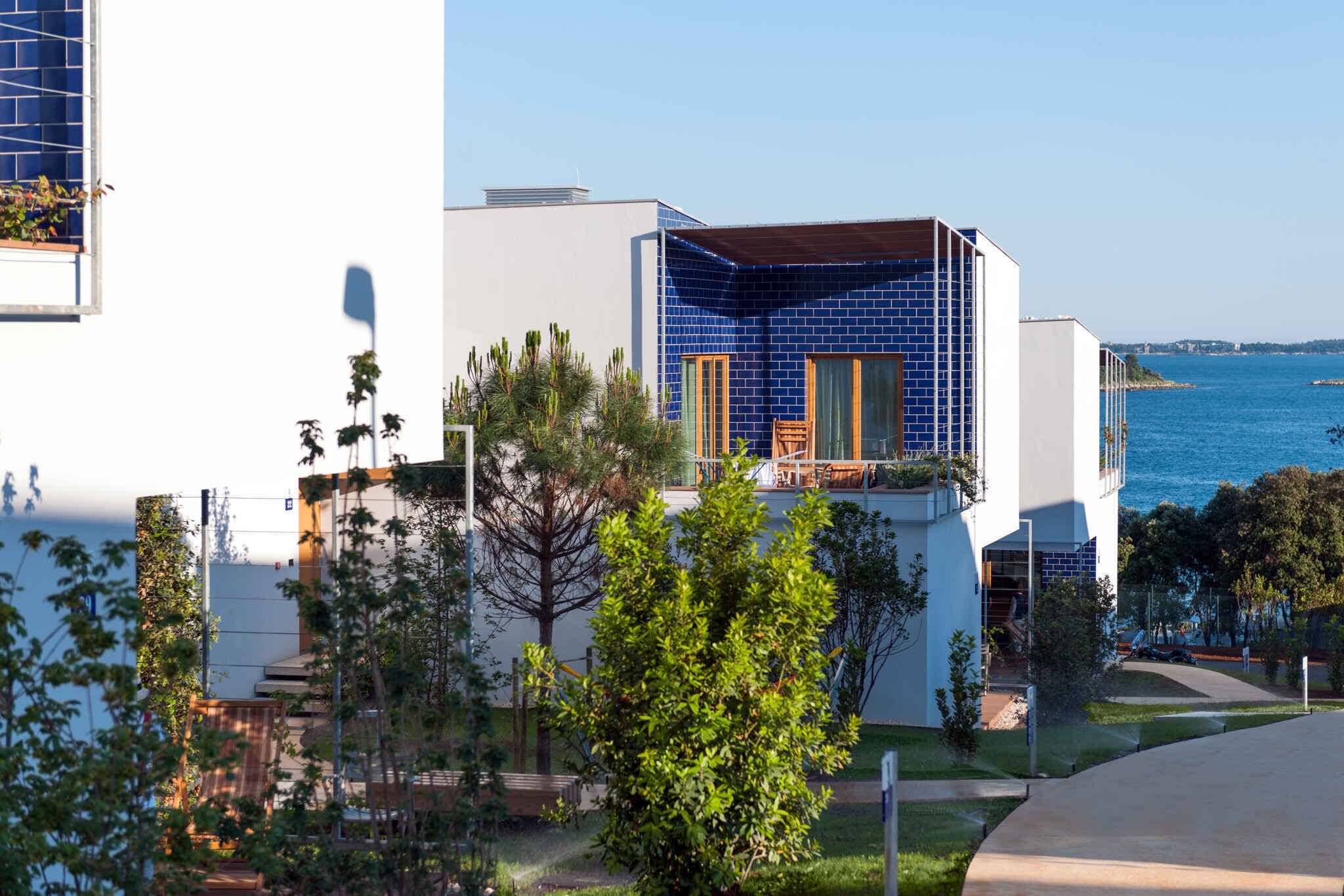 A modern building with a blue-tiled corner, surrounded by greenery and overlooking a body of water.