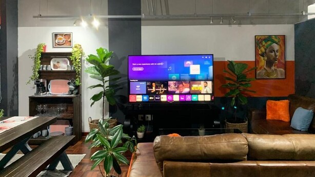 Una sala moderna con un televisor de pantalla plana, un sofá de cuero marrón y una mesa de comedor con sillas. Hay plantas en macetas y un cuadro en la pared.