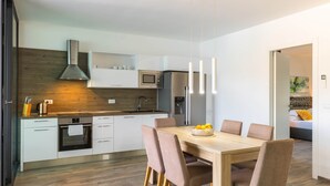 Ett modernt kök med vita skåp, en träpanel bakom arbetsbänken och rostfria köksmaskiner. Det finns en matplats med ett träbord och stolar.