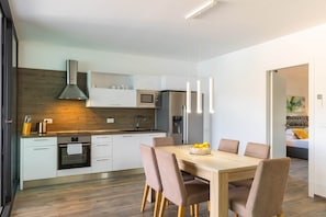 Una cocina moderna con armarios blancos, un salpicadero de madera y electrodomésticos de acero inoxidable. Hay un comedor con una mesa y sillas de madera.