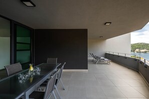 A modern outdoor dining area with a glass table and chairs, a dark wall, and a view of a body of water and greenery. - Korcula Luxury Apartments - Olive 6 (Žrnovo)