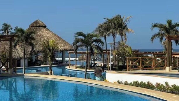 Ein Poolbereich eines Resorts mit einer strohgedeckten Struktur, Palmen und Blick auf den Ozean.