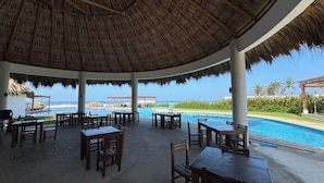 Ein überdachter Außenbereich mit Tischen und Stühlen, mit Blick auf einen Pool und einen Strand.