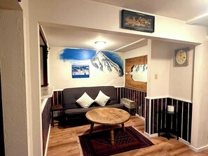 Ein Zimmer mit einer Couch, einem runden Holztisch, einem roten Teppich, einem gerahmten Bild an der Wand und einer kleinen Holzkabine.