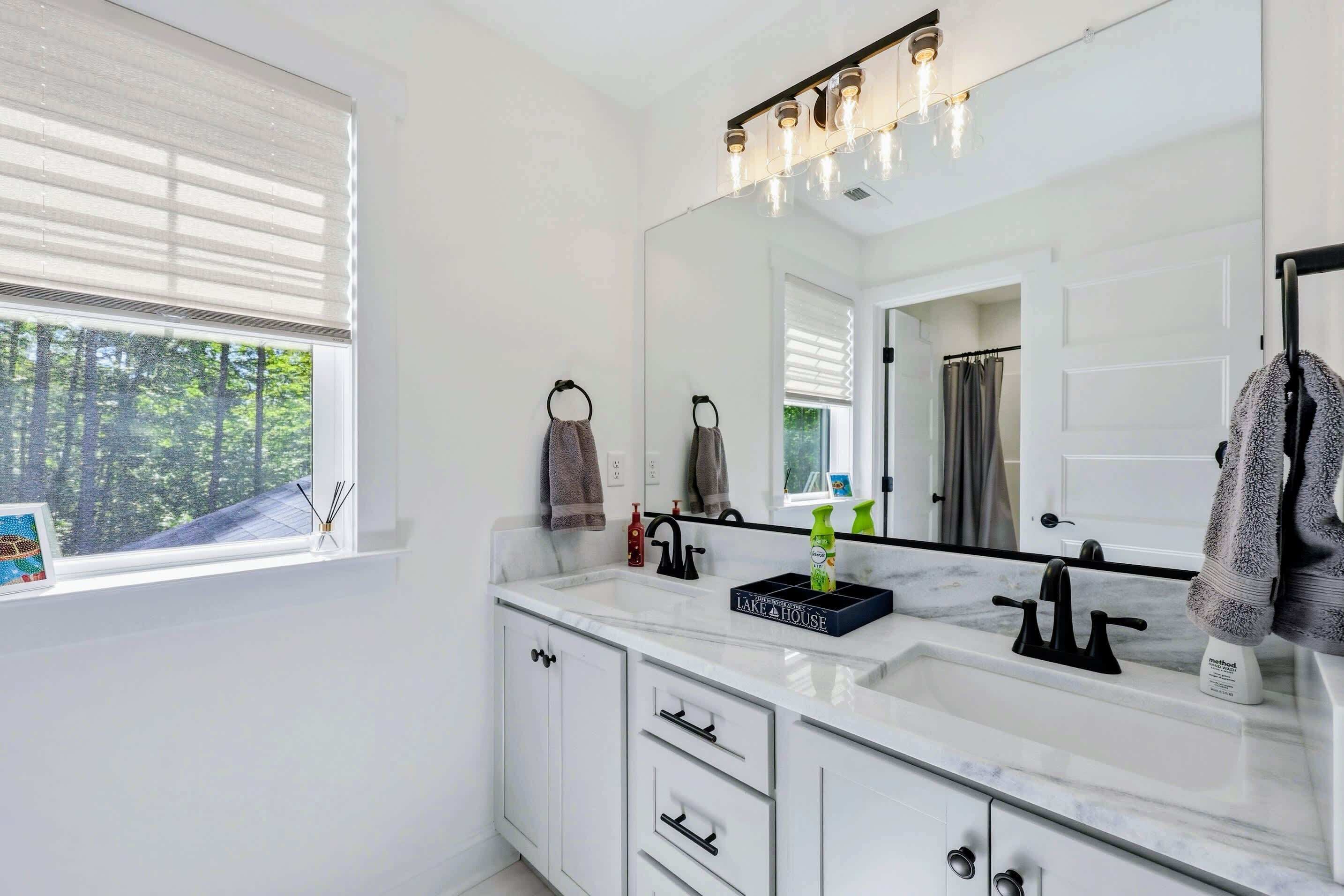 Un baño con un espejo grande, muebles blancos, un lavabo y una ventana con persianas.