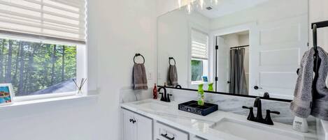 Un baño con un espejo grande, muebles blancos, un lavabo y una ventana con persianas.