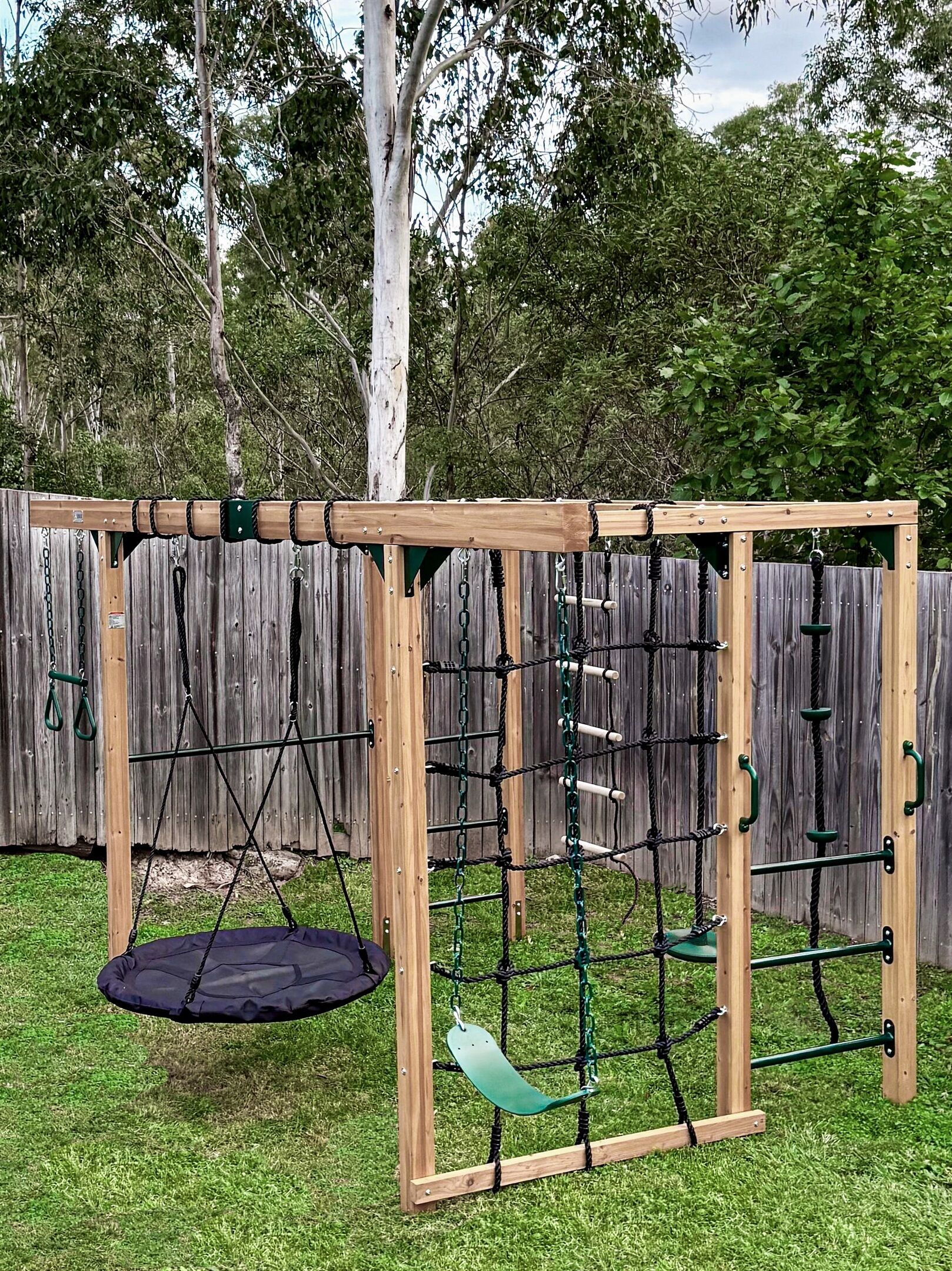 A wooden playground structure with a swing, climbing net, and a slide.
