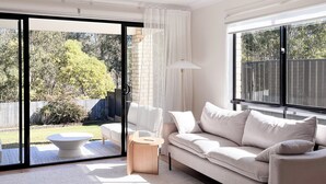 Un salon moderne avec un canapé, une table basse ronde et un lampadaire. Une porte-fenêtre coulissante donne accès à un jardin agrémenté d’une clôture en bois et d’arbres.