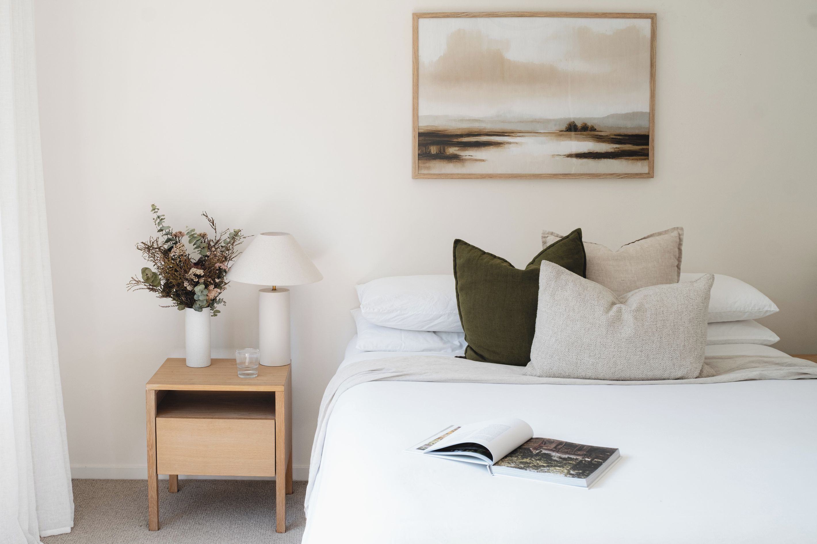 A neatly arranged bedroom with a bed, pillows, a bedside table with a lamp, a vase with dried flowers, and a framed landscape painting on the wall.