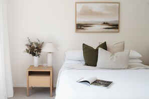 A neatly arranged bedroom with a bed, pillows, a bedside table with a lamp, a vase with dried flowers, and a framed landscape painting on the wall.