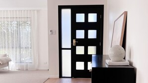 Une pièce avec une porte noire, un canapé blanc, une grande fenêtre avec des rideaux légers et un objet sphérique blanc posé sur un support noir.