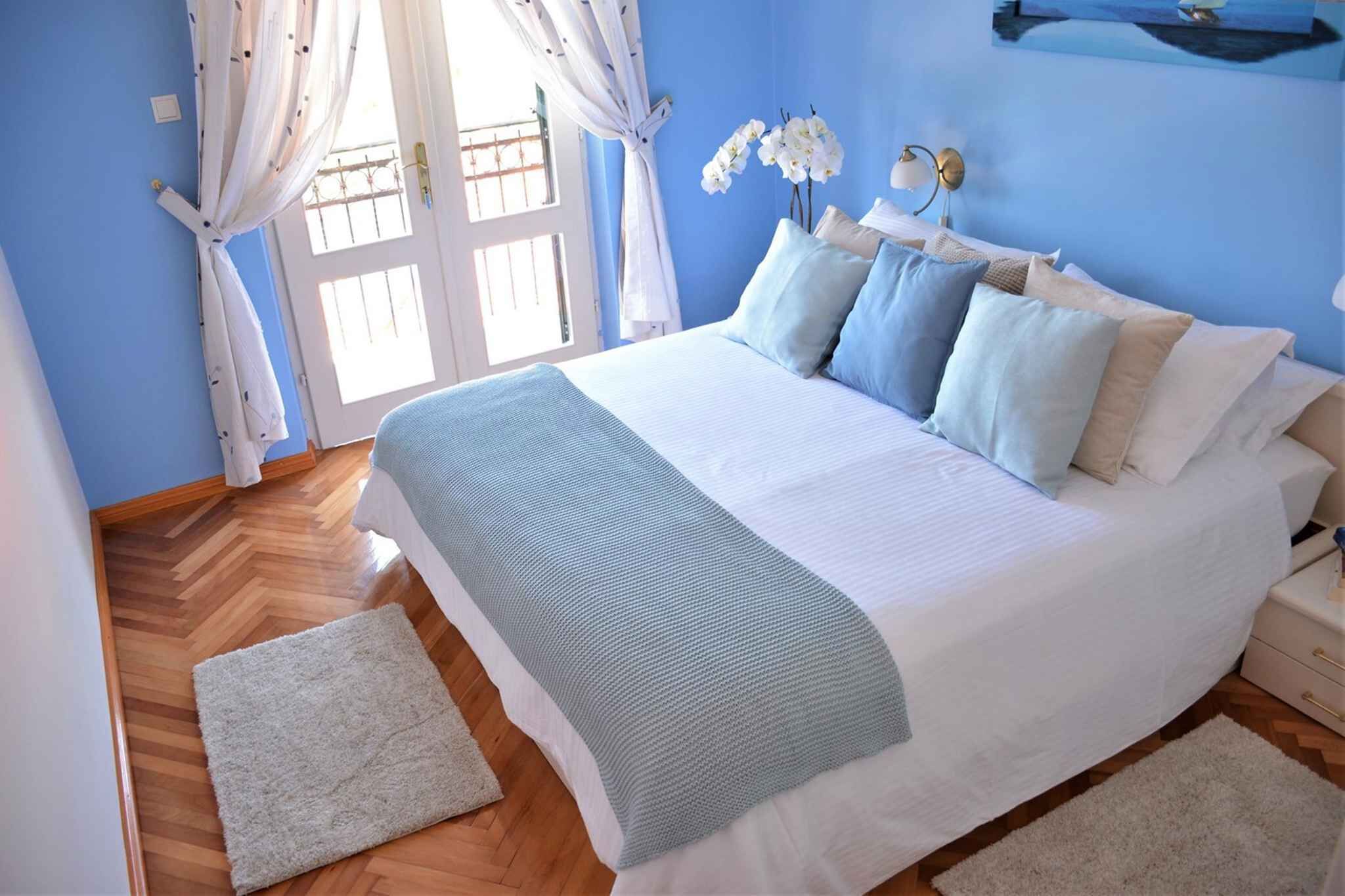 A bedroom with a bed, white curtains, a window with a view, and a wall-mounted light fixture.