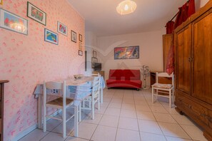 A room with a dining table, chairs, a red sofa, a wooden cabinet, and a ceiling fan. - APPARTAMENTI LENTISCO - BILOCALE LENTISCO (Capoliveri)