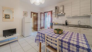 A compact kitchen with white cabinets, a dining table with a checkered tablecloth, a television on a stand, and a refrigerator.