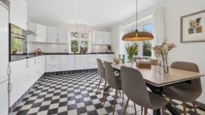 Uma cozinha moderna com piso xadrez, armários brancos, uma mesa de jantar com cadeiras e iluminação pendente.