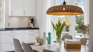 Uma cozinha com armários brancos, uma luminária de vime suspensa, um vaso com tulipas e uma mesa de jantar com cadeiras.