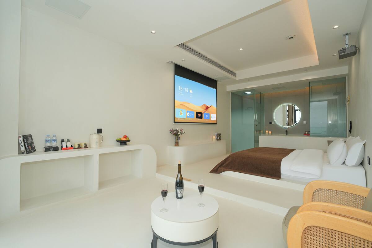 Una habitación de hotel moderna con una cama, una zona de estar con una mesa, un televisor montado en la pared y un espejo grande.