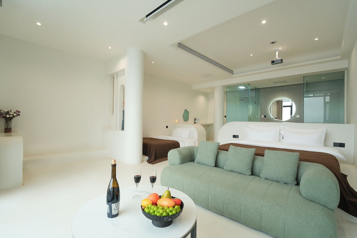 Una habitación de hotel moderna con un sofá verde, una mesa redonda con una botella y fruta, y una cama con ropa blanca.