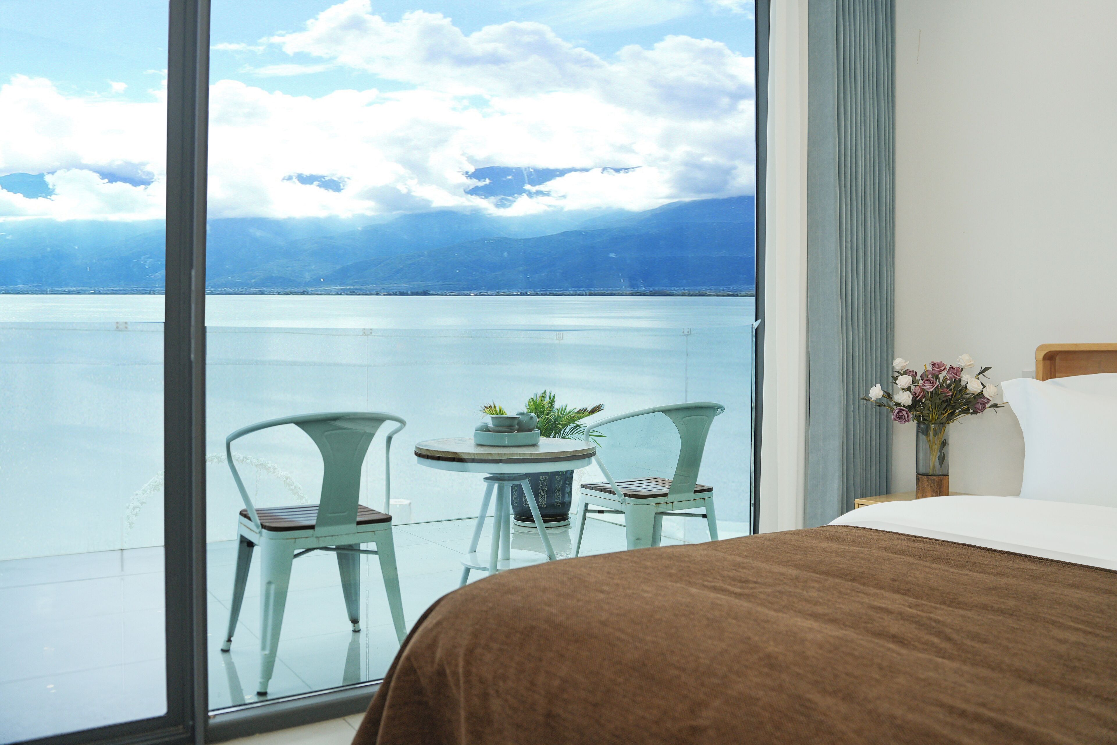A hotel room with a large window overlooking a body of water and mountains. There is a bed with a brown cover, two chairs, and a small table with a plant.