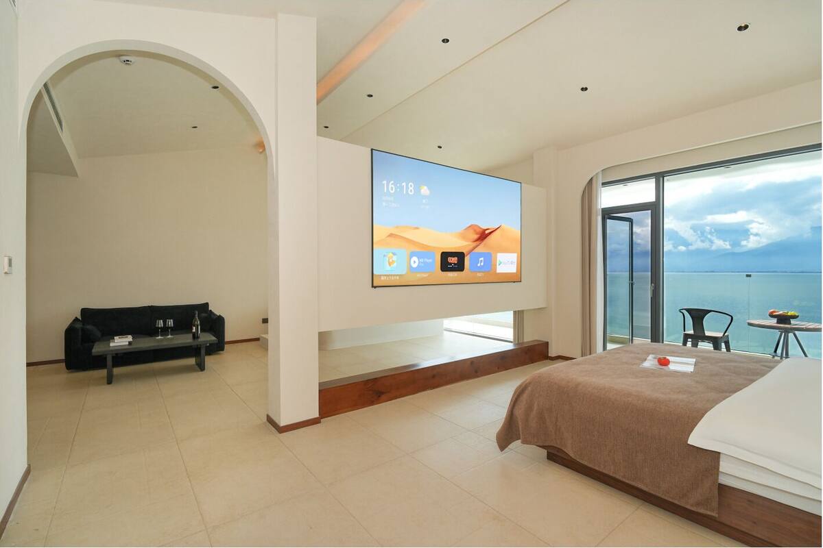 Habitación de hotel moderna con un televisor grande, una cama, un sofá y un balcón con vistas al mar.