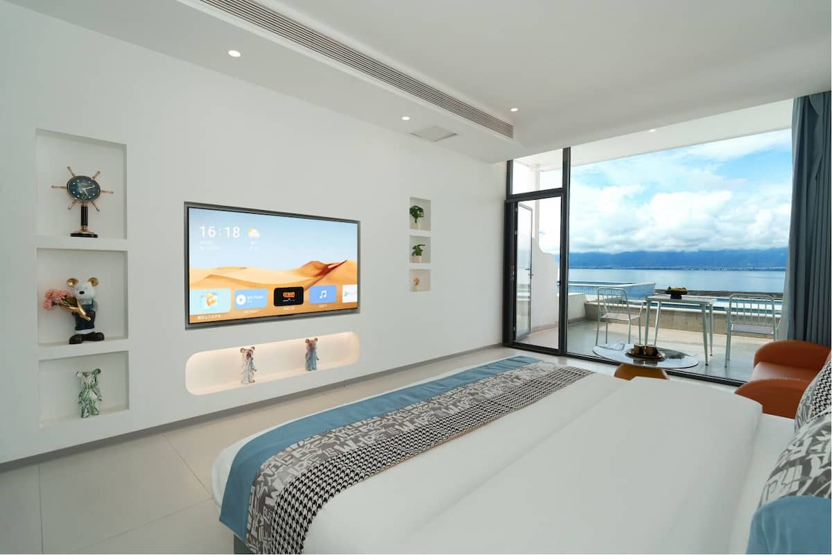Un dormitorio moderno con un televisor grande, una cama con ropa de cama estampada, un balcón con vistas al mar y elementos decorativos en las paredes.