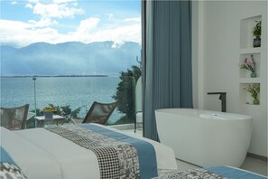 A bedroom with a large window overlooking a lake and mountains, a white bathtub, and a small table with fruit. - Guiyue Mountain Seaview Hotel (Dali)