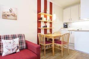 A compact living space with a red sofa, a wooden dining table with four chairs, a kitchen area with white cabinets, and a striped wall decoration. - Apartment im Seepark Sellin mit Balkon (Sellin)