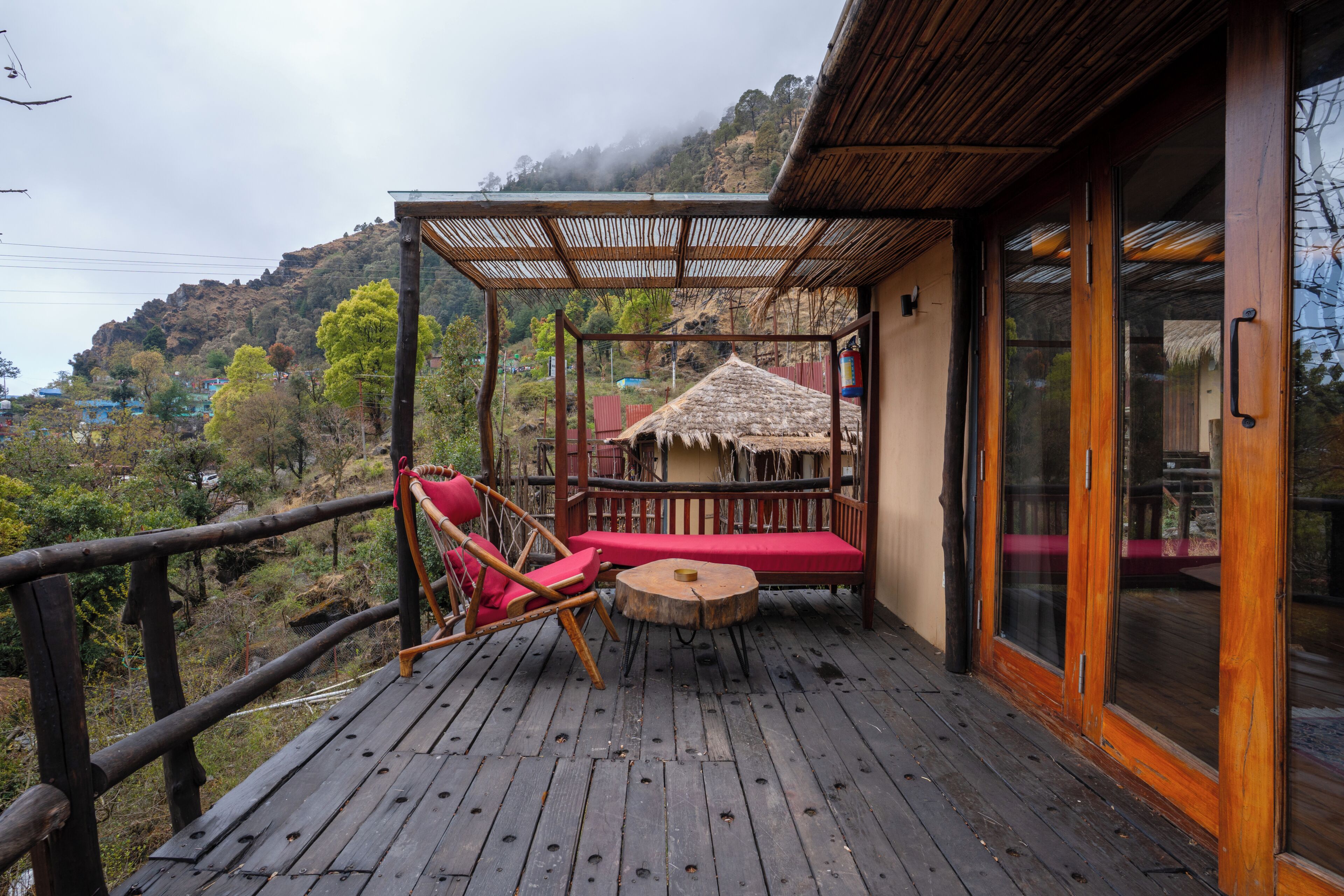 A wooden deck with a red cushioned seating area, a small round table, and a thatched-roof structure in the background.