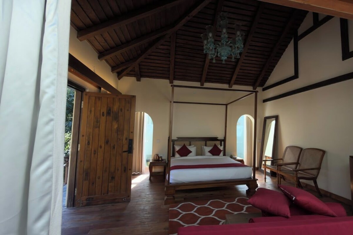 A bedroom with a large bed, wooden ceiling beams, a chandelier, and a view through a window.