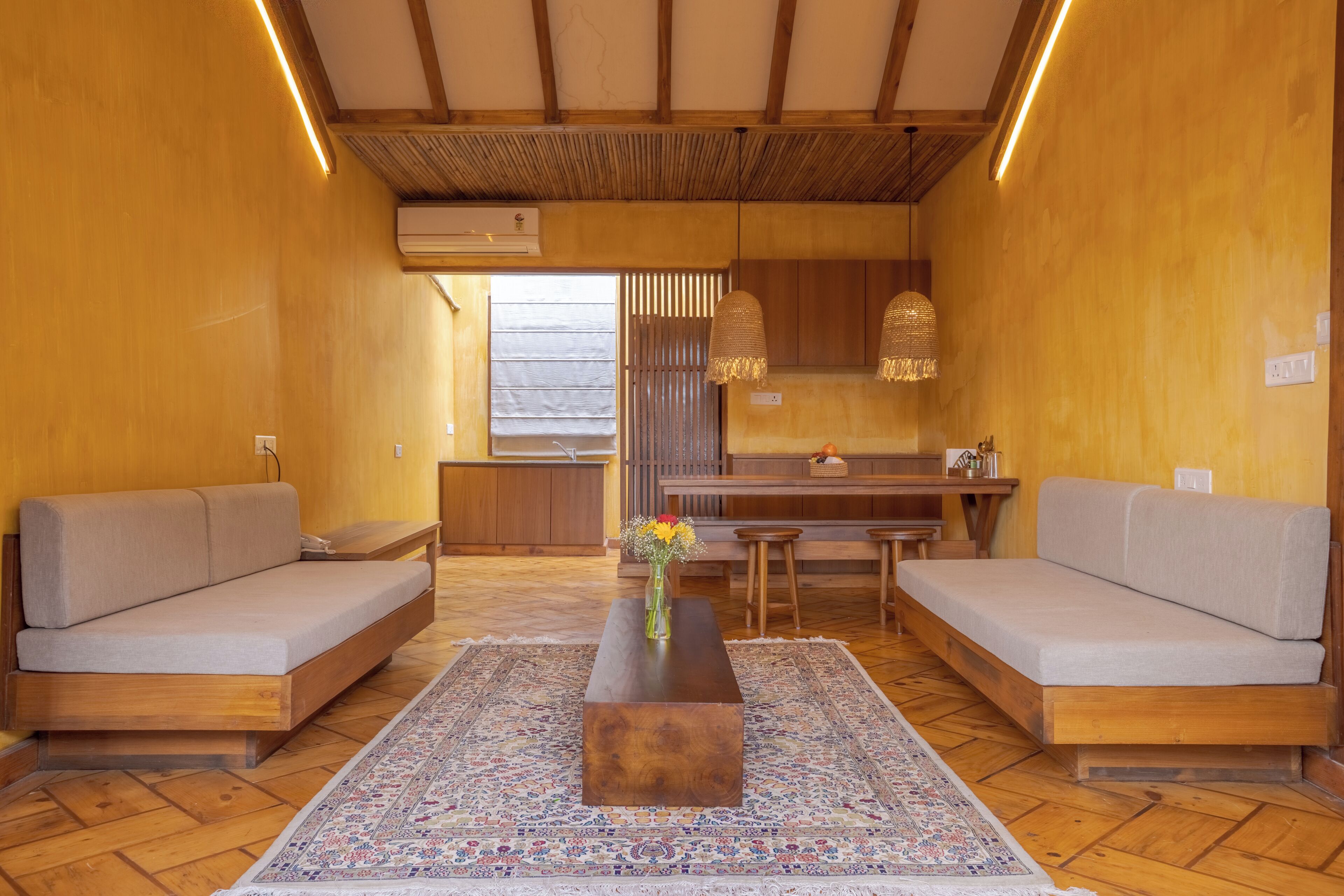Une pièce comprenant deux canapés, une table basse en bois, un tapis à motifs et un coin cuisine avec des meubles en bois et un luminaire suspendu.