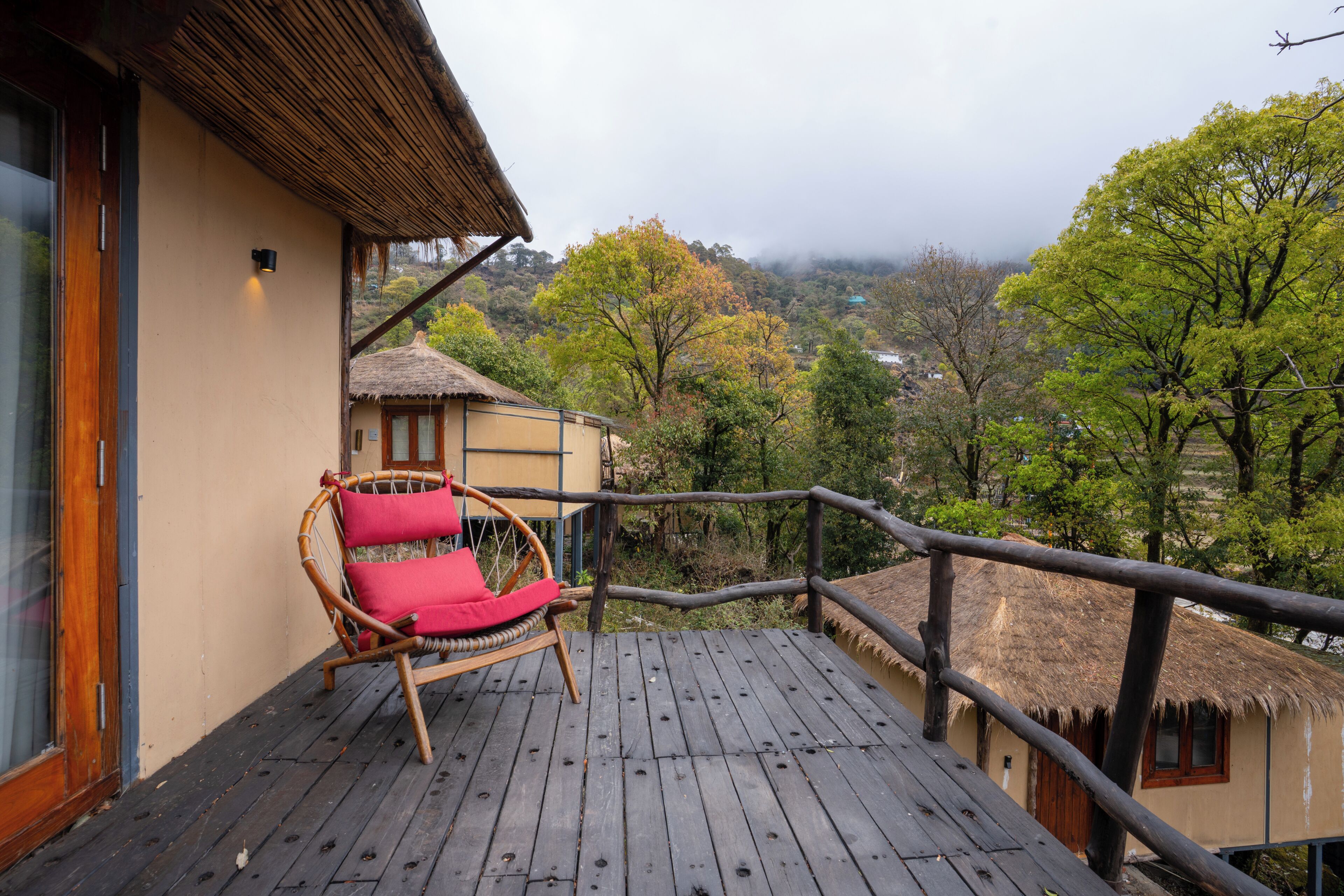 Una terraza de madera con una silla de mimbre, un edificio con techo de paja y vistas a un paisaje boscoso.