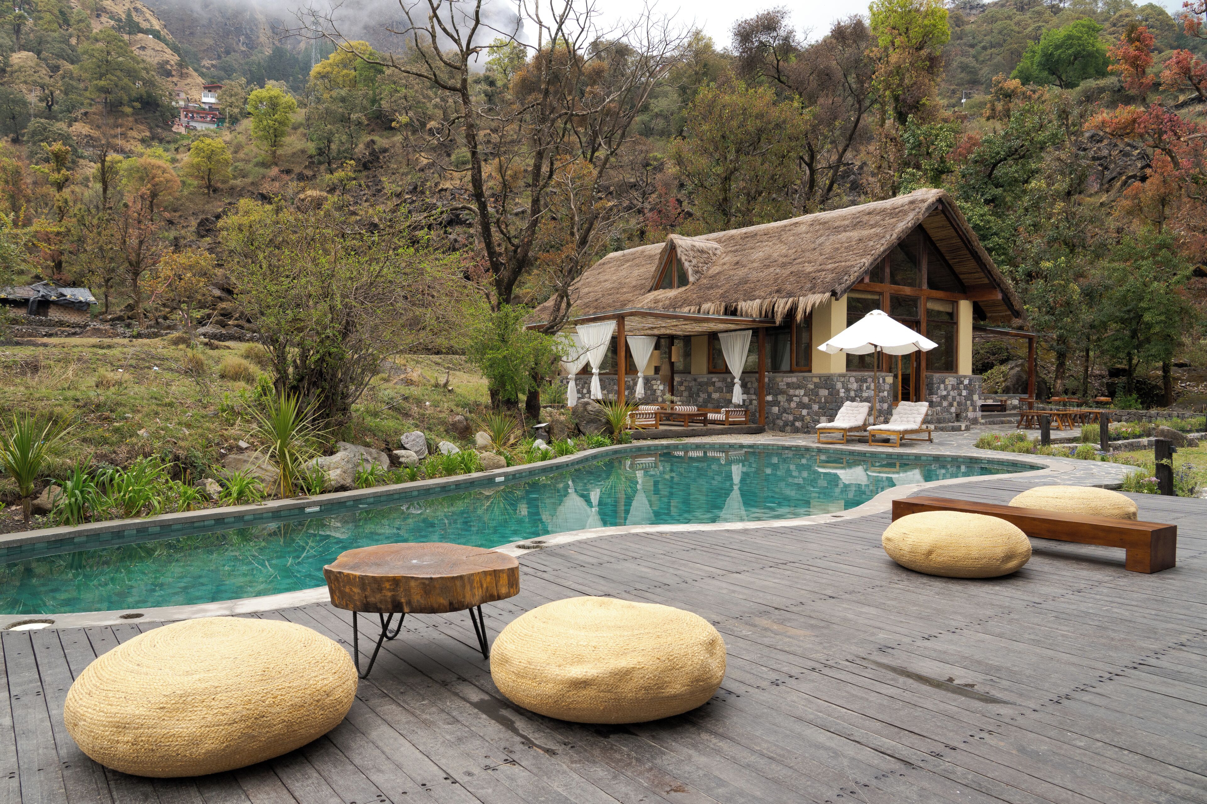 Un área de piscina con terraza de madera, sillas de descanso y un edificio con techo de paja.