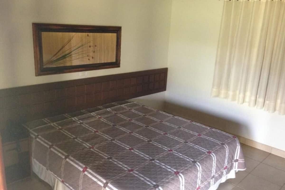 Un dormitorio con cama, cabecera, un cuadro enmarcado en la pared y ventana con cortinas.