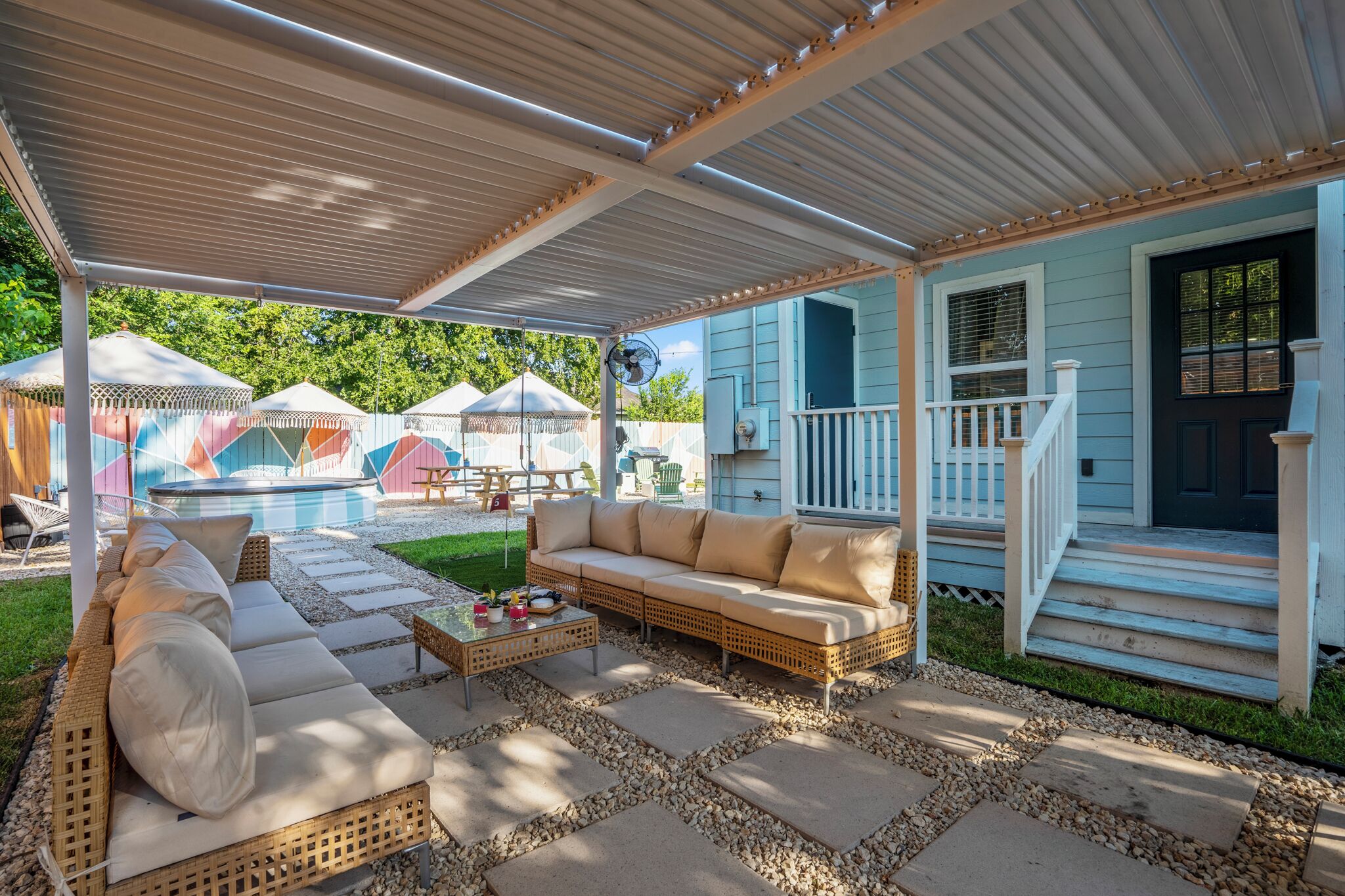 A covered outdoor seating area with a wicker sofa and a coffee table, a wooden deck, and a house with a blue exterior and white trim.