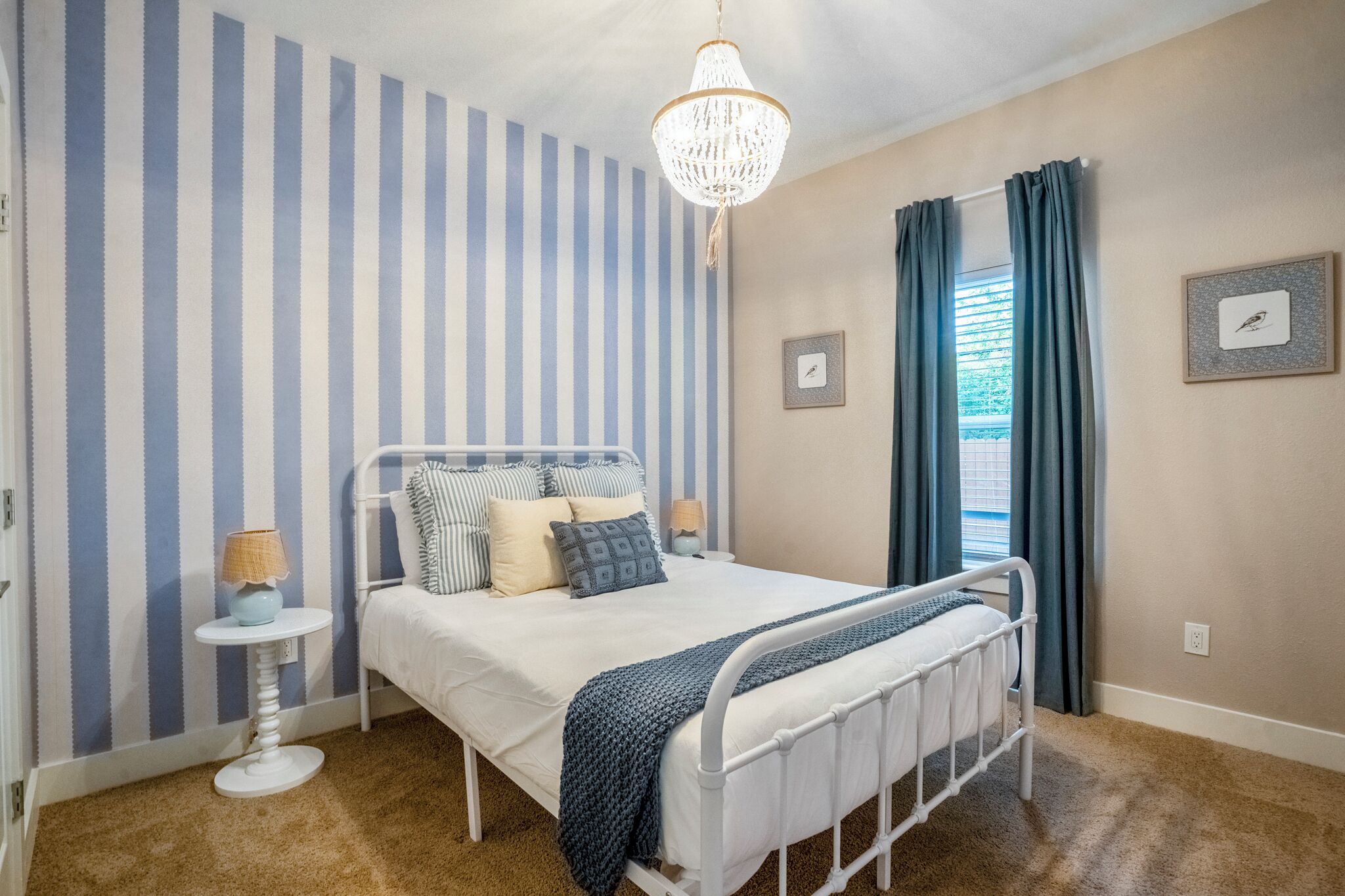 A bedroom with a striped wall, a bed with pillows, a bedside lamp, a chandelier, a window with curtains, and a framed picture on the wall.