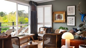 A cozy living room with wicker furniture, a large window with blinds, and framed botanical prints on the wall. - Bengarlyn in Daylesford where bold garden design meets boutique luxury, stunning 3 bedroom home (Daylesford)