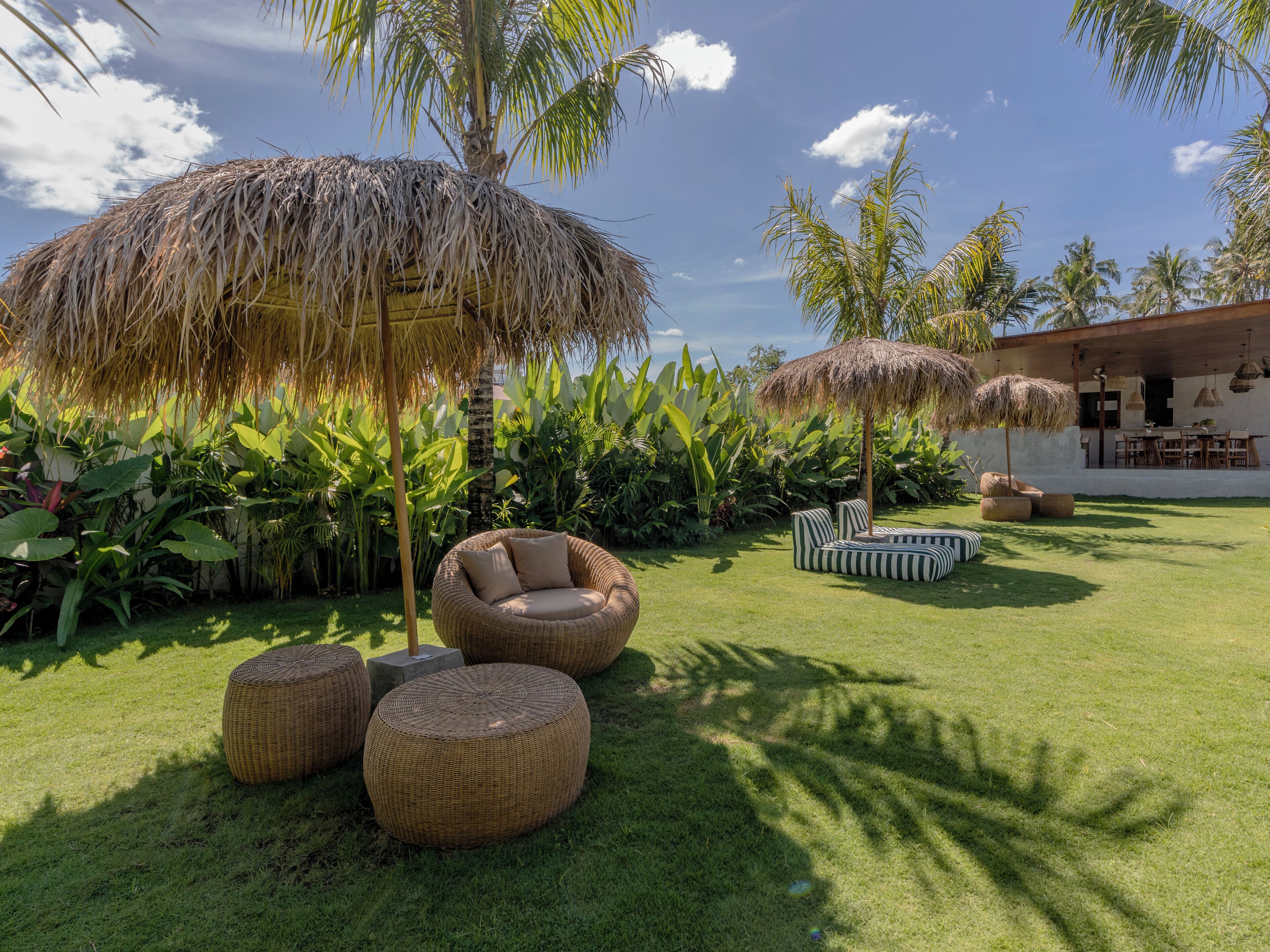 Un espace extérieur aménagé dans un style tropical, avec des meubles en osier et des parasols en chaume, entouré d’une végétation luxuriante et de palmiers.