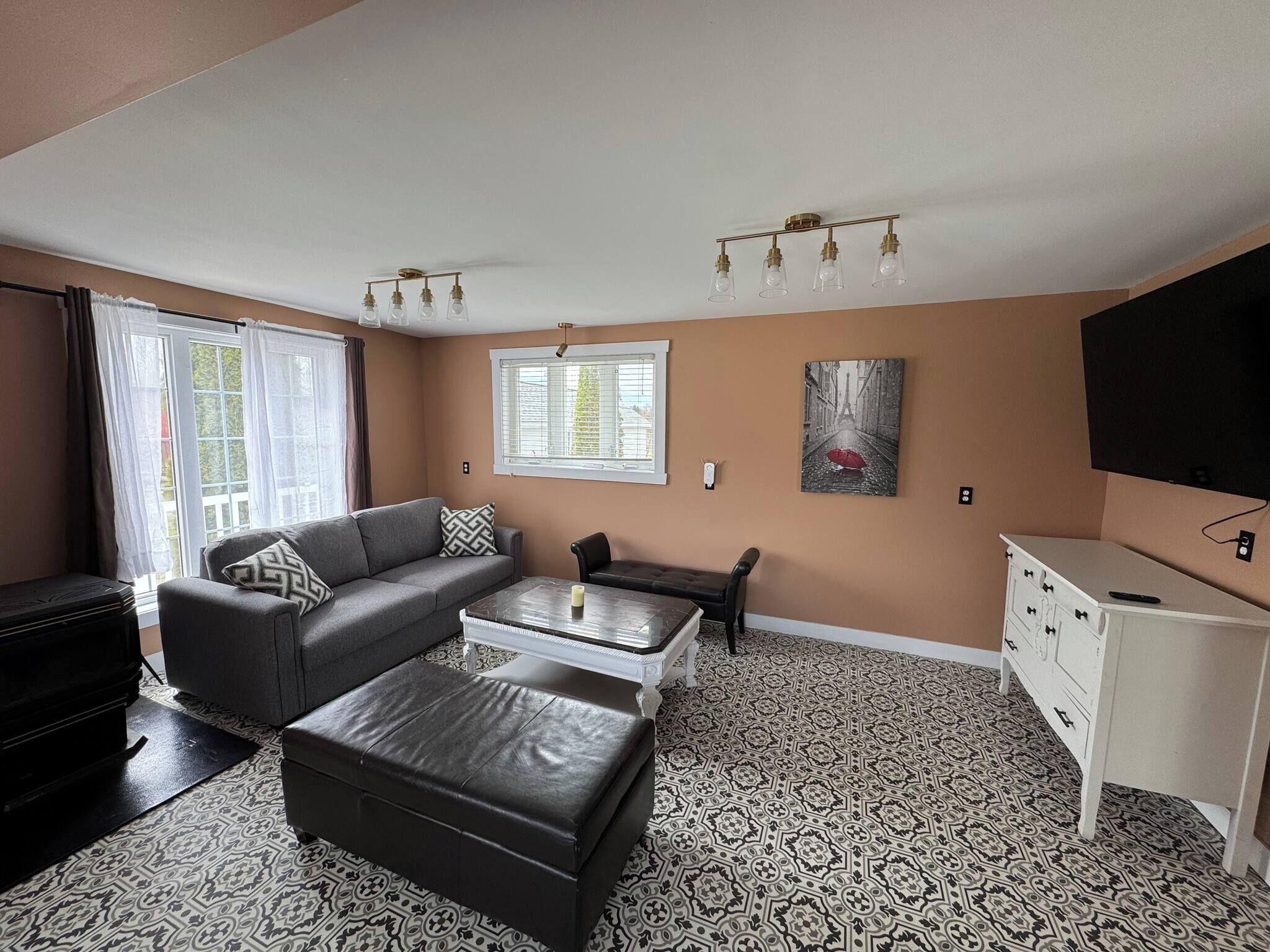A living room with a sofa, ottoman, and a coffee table. There is a window with blinds, a painting on the wall, and a television mounted on the wall.