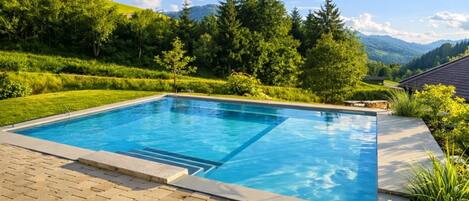 A rectangular swimming pool with a paved deck, surrounded by lush greenery and hills in the background.