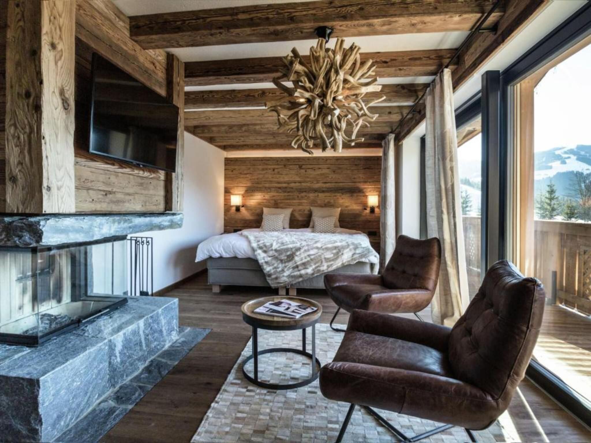 A modern bedroom with a large bed, a fireplace, a leather armchair, and a view of the mountains.